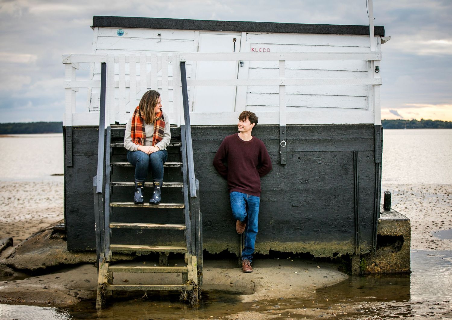 Two people sit together on wooden steps outside a white building near the water.