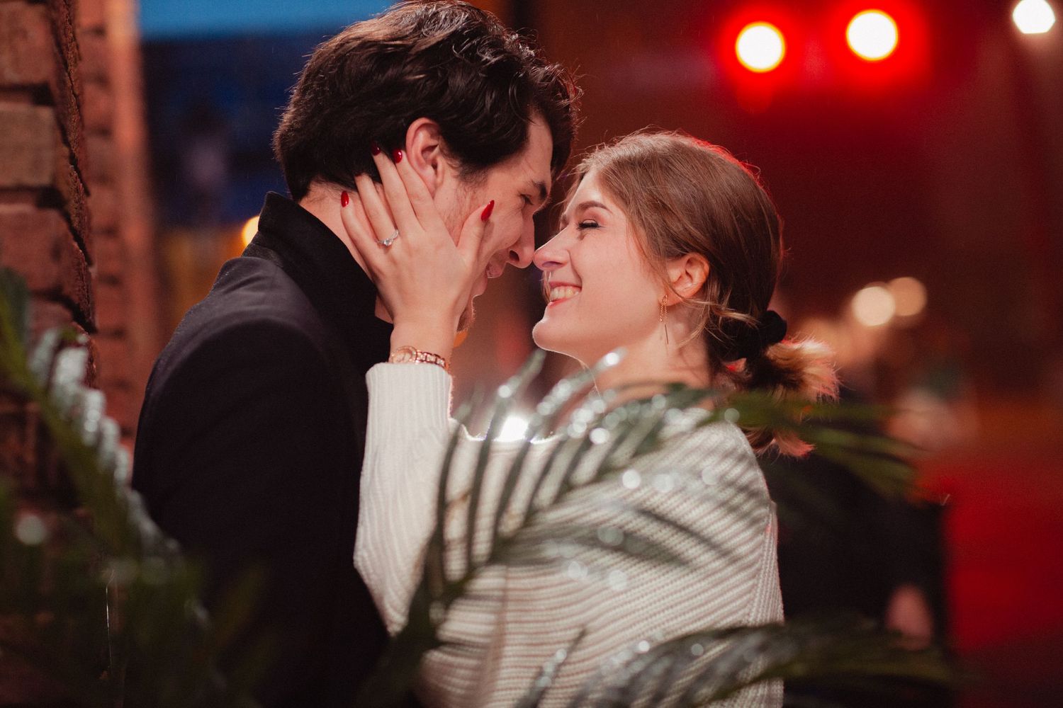 Two people share a tender moment under dramatic red lighting with greenery visible.