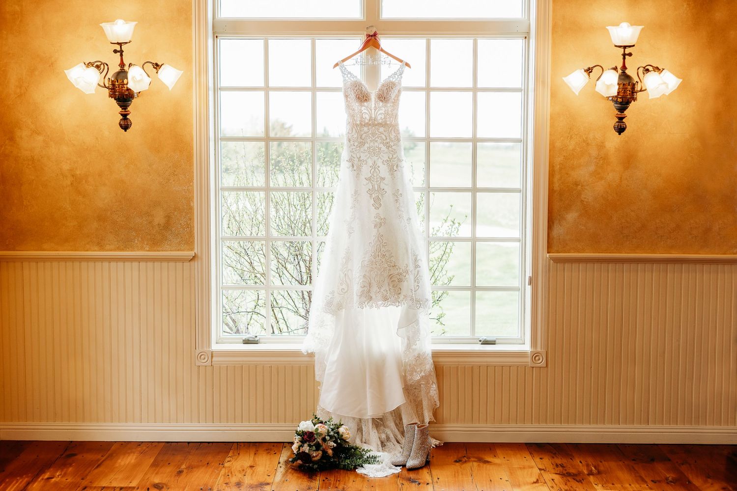 A white lace wedding dress hangs elegantly in front of a bright window with ornate wall sconces on either side.