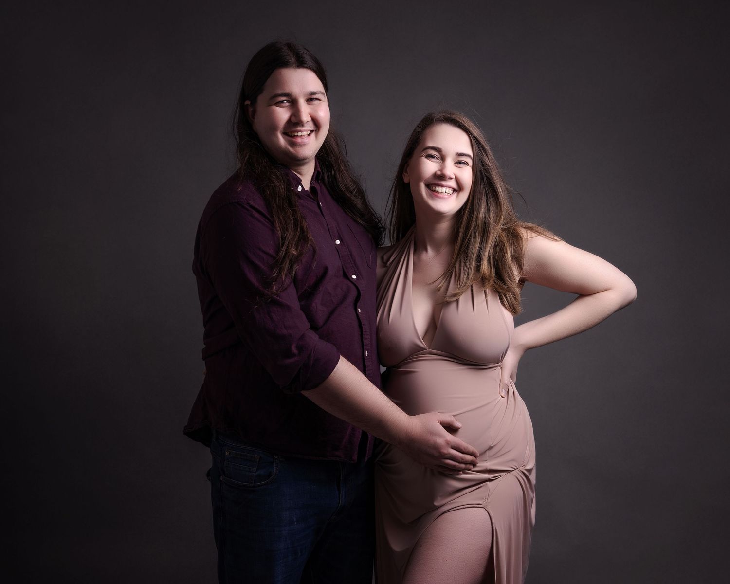 Two smiling figures pose together for a maternity portrait against dark background.