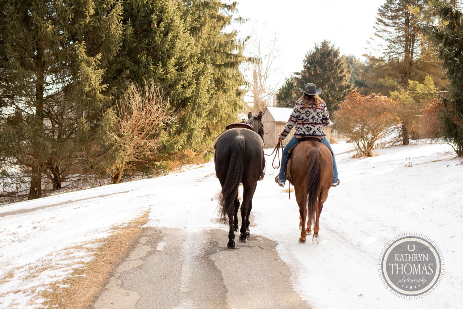 Two riders on horseback travel down a snowy path surrounded by winter trees.