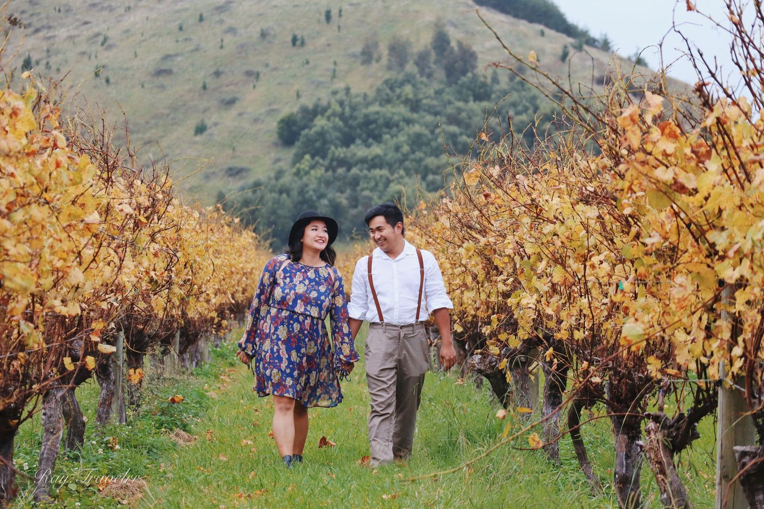 A couple walking through golden autumn vineyard rows with mountains in the background.