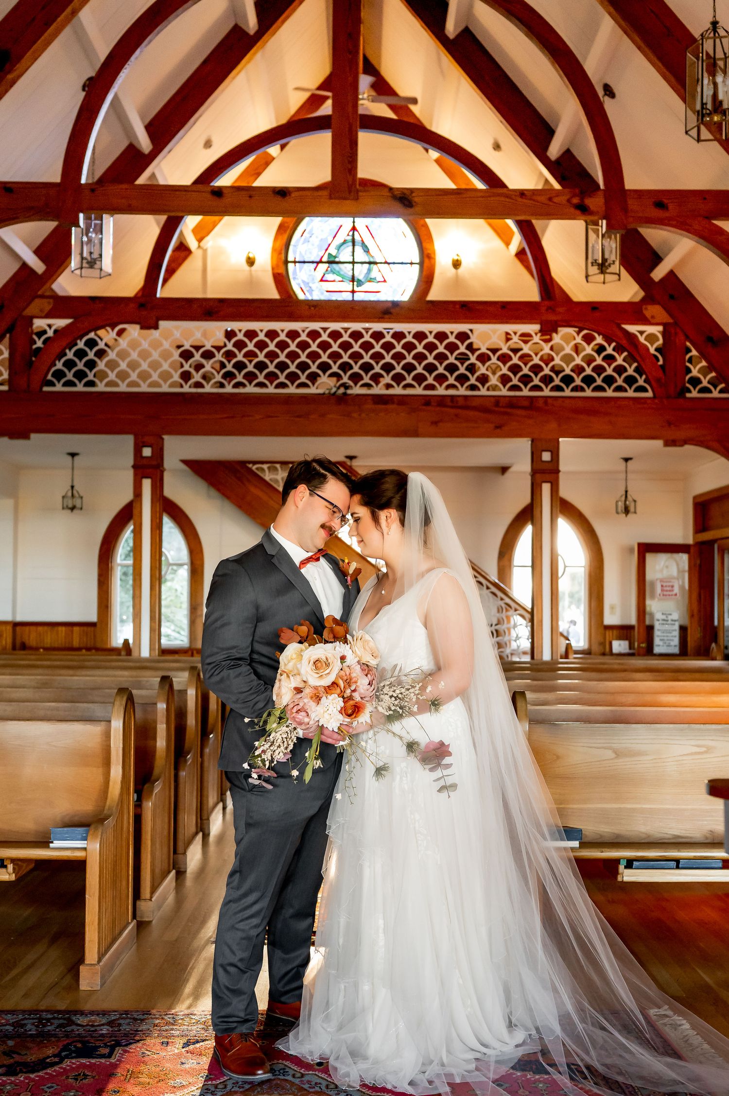 A wedding couple shares an intimate moment in a chapel with dramatic wooden beam architecture.