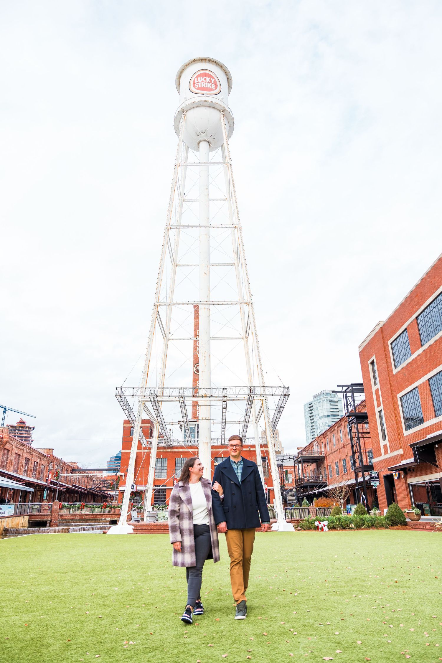 Couple walks hand in hand on a green lawn in front of a tall historic water tower at a brick urban venue.