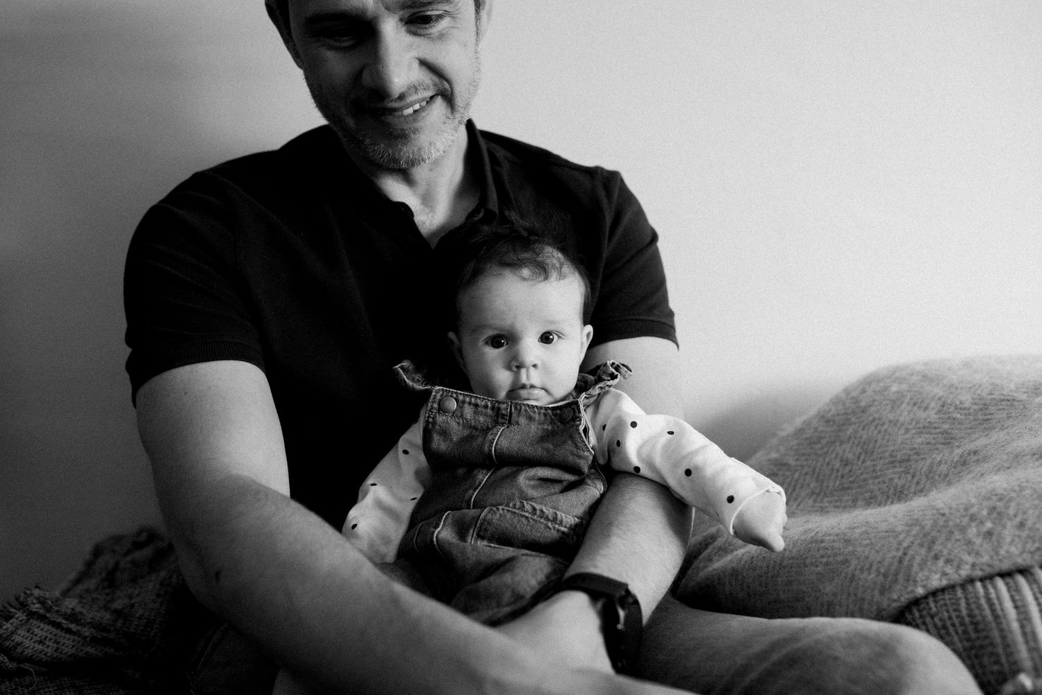 Black and white photo of a baby in overalls being held on a couch.