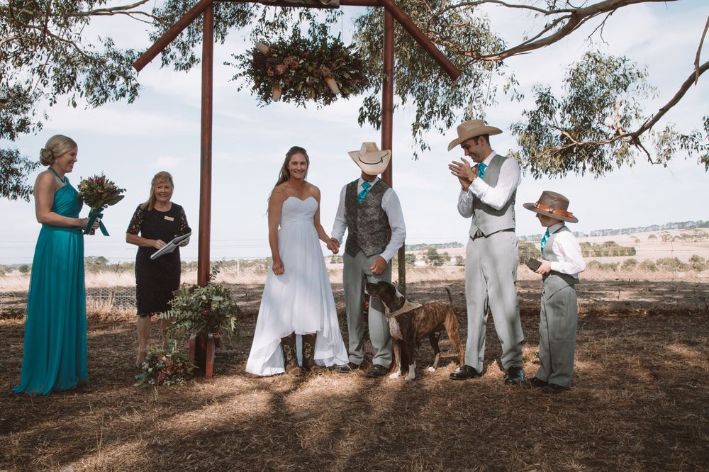 Rustic outdoor wedding ceremony under wooden arch with native trees in background.