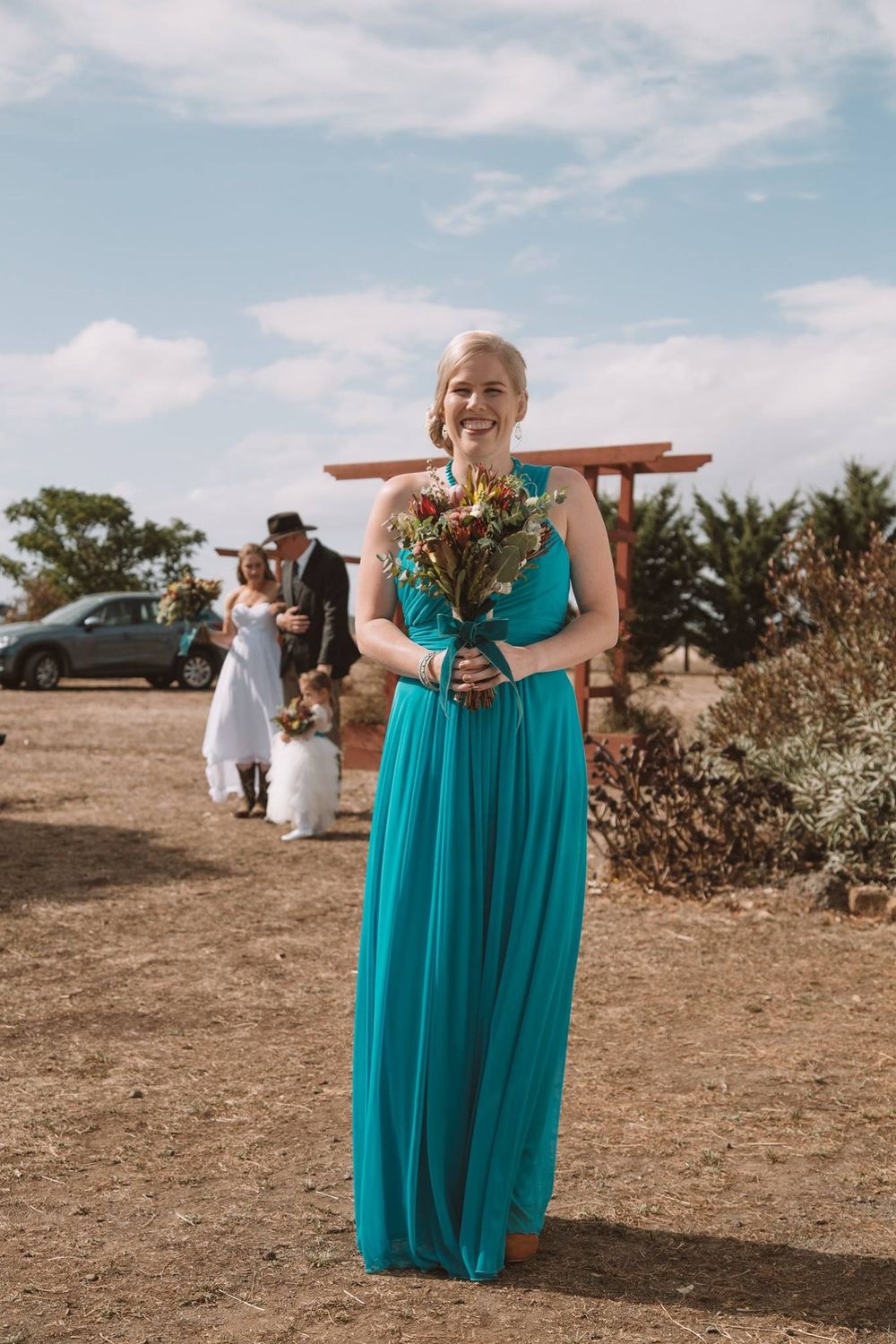 Person in turquoise dress holds bouquet at outdoor wedding with wooden arch in background.