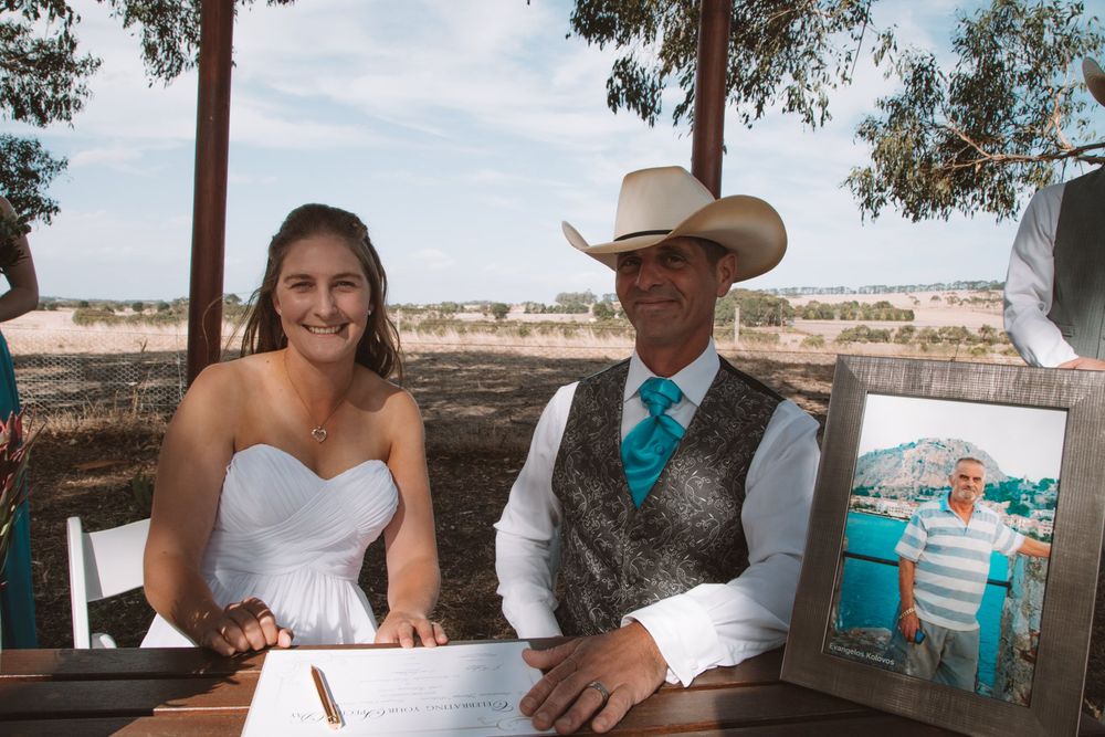 Couple in western wedding attire sits at outdoor table signing documents.