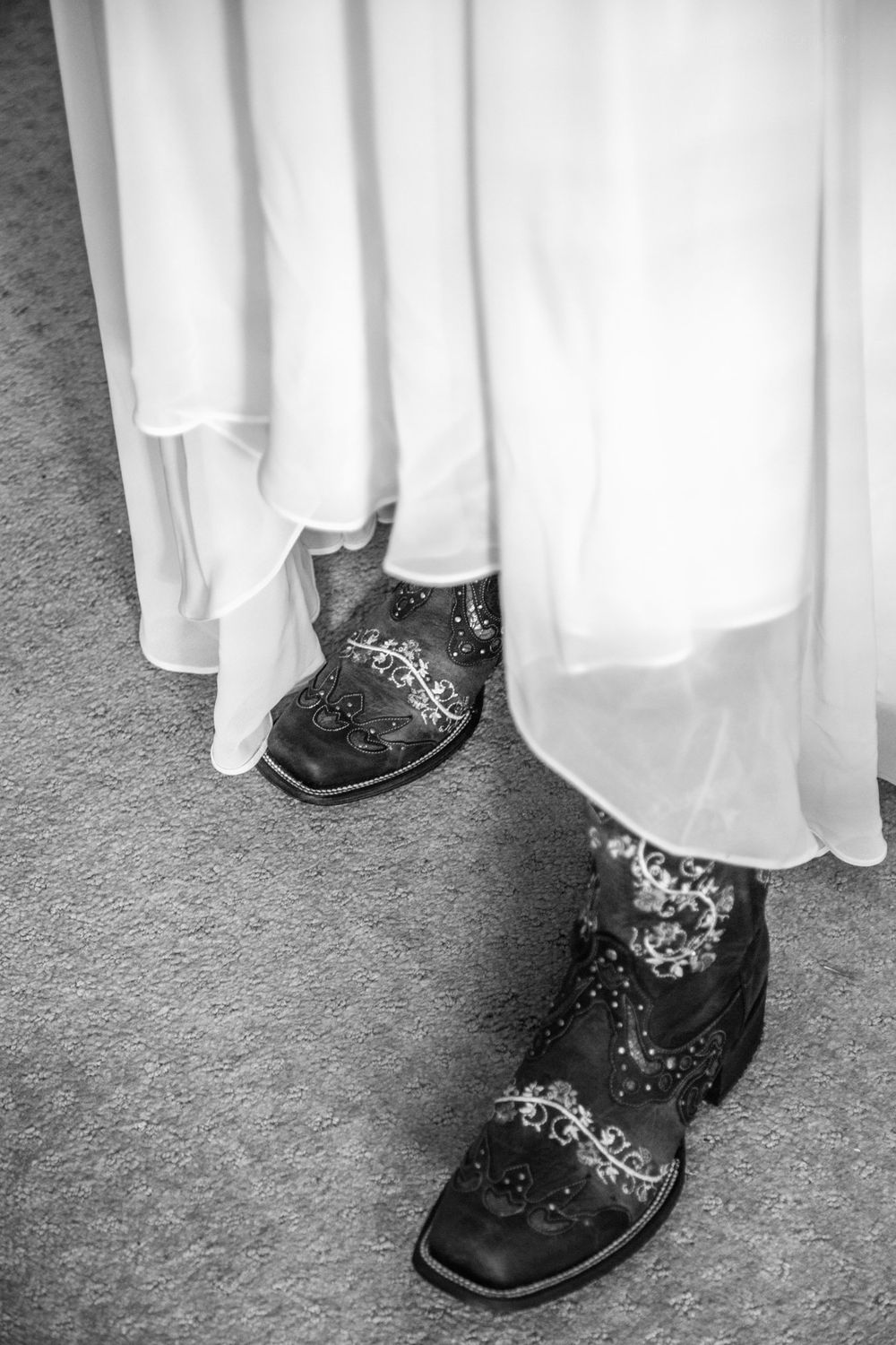 Close-up of embroidered black boots peeking out from under a white dress against a gray carpet.