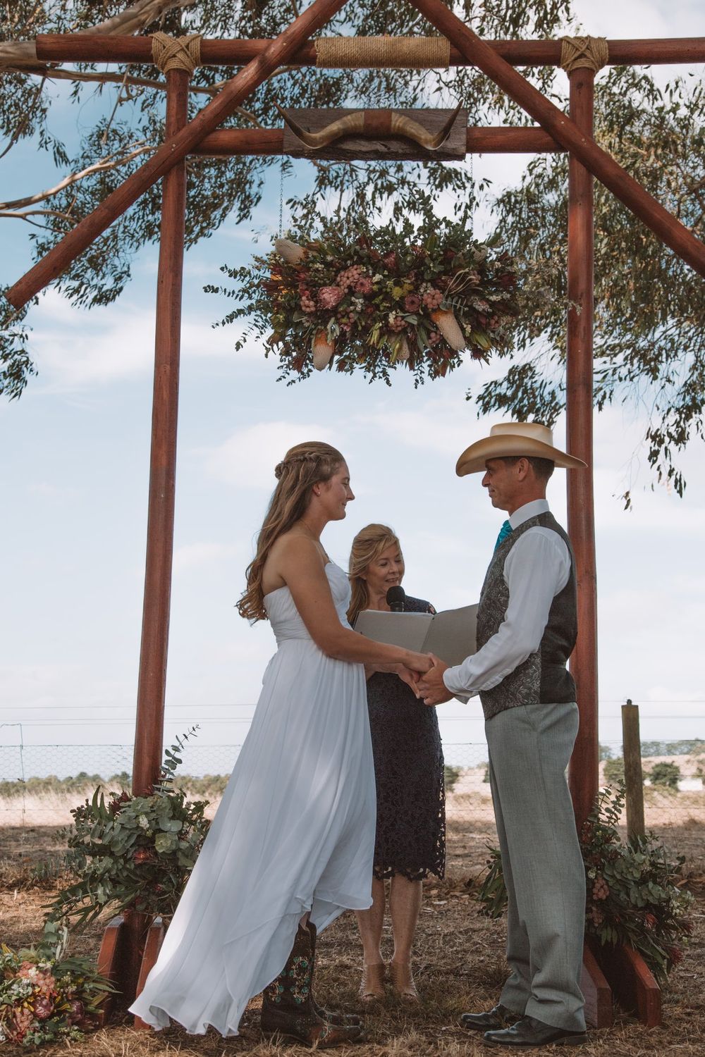 Wedding ceremony under wooden arch with suspended floral arrangement.