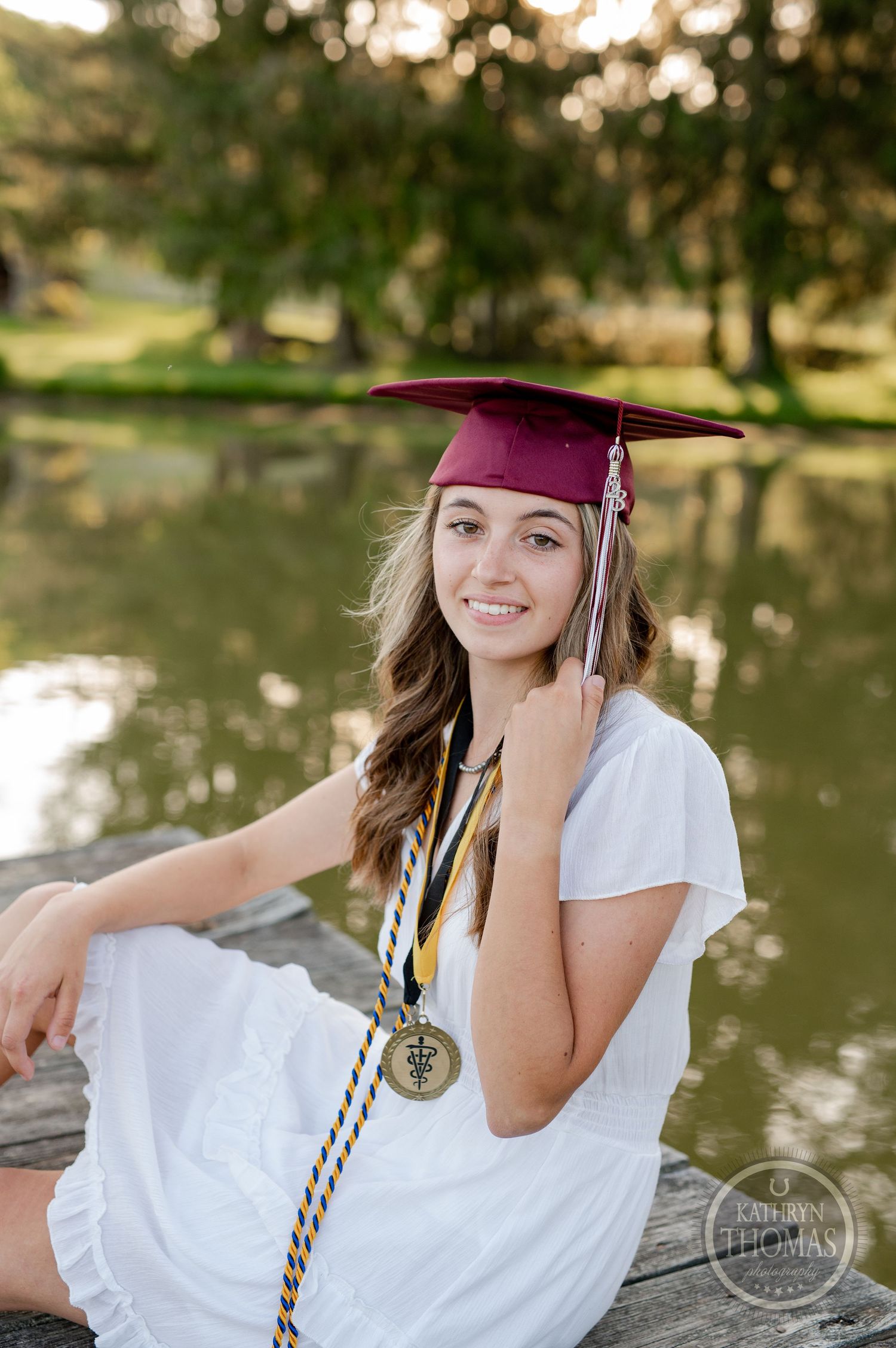 Graduate in burgundy cap poses with medal while sitting on dock by pond.