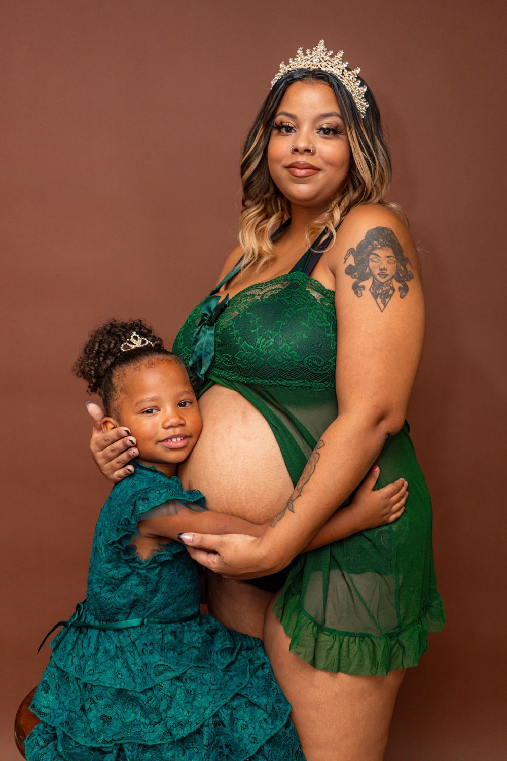 Pregnant mom wearing crown and green lingerie embraces young daughter in matching teal dress.