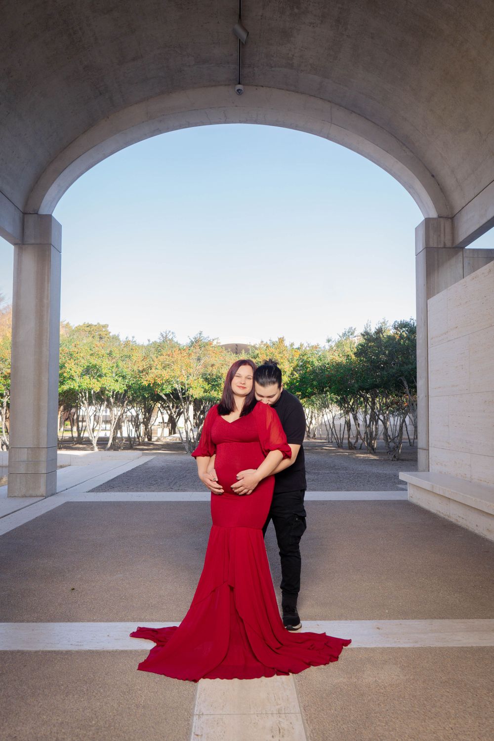 Maternity couple photo under stone arch, woman in red gown with train embraced by partner.