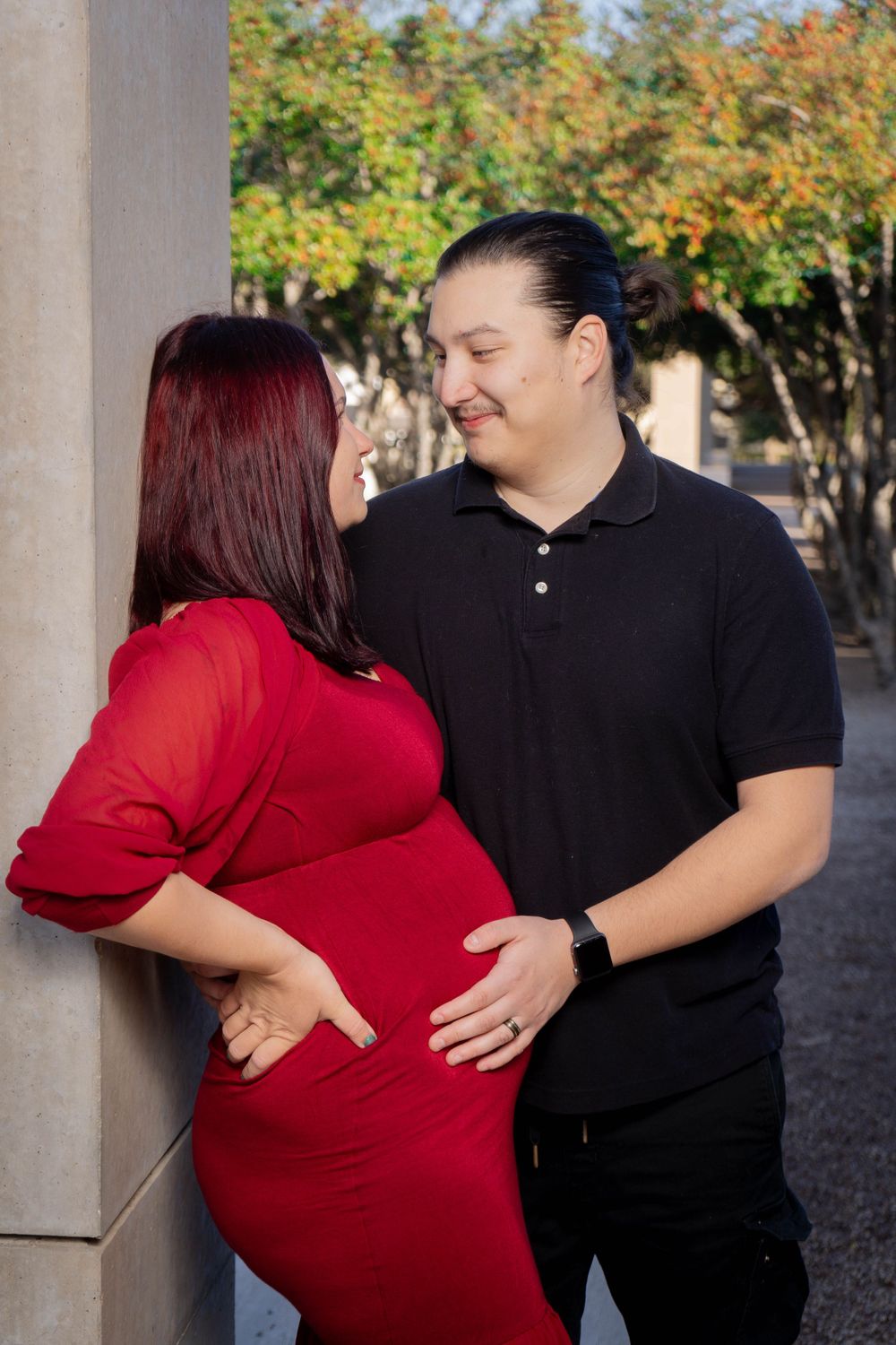 Expecting couple in maternity photo, woman in red dress and man in black shirt outdoors.