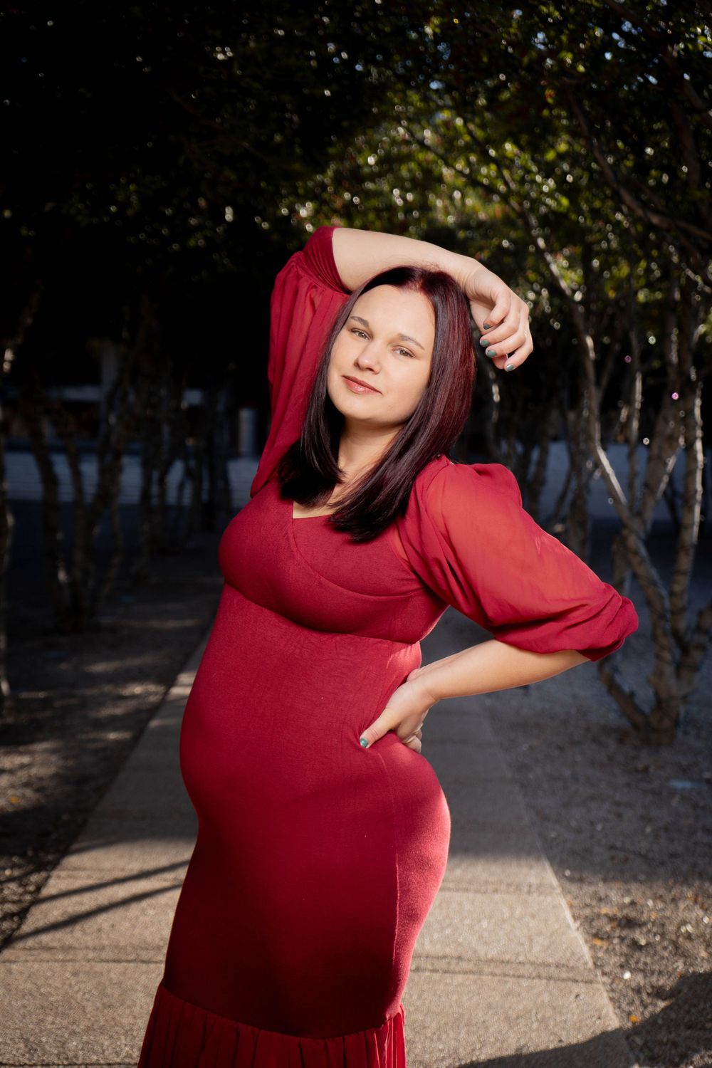 Pregnant woman in red dress posing outdoors among trees in maternity photo.