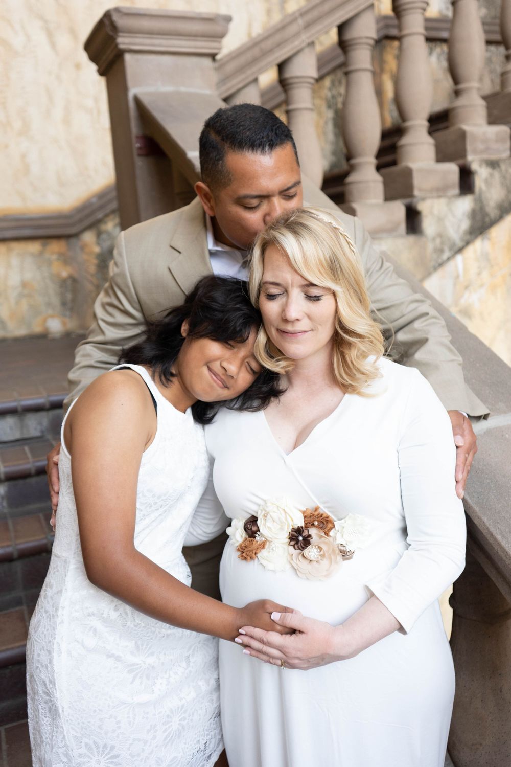 Pregnant woman in white floral dress embraced by husband and daughter on stone staircase.
