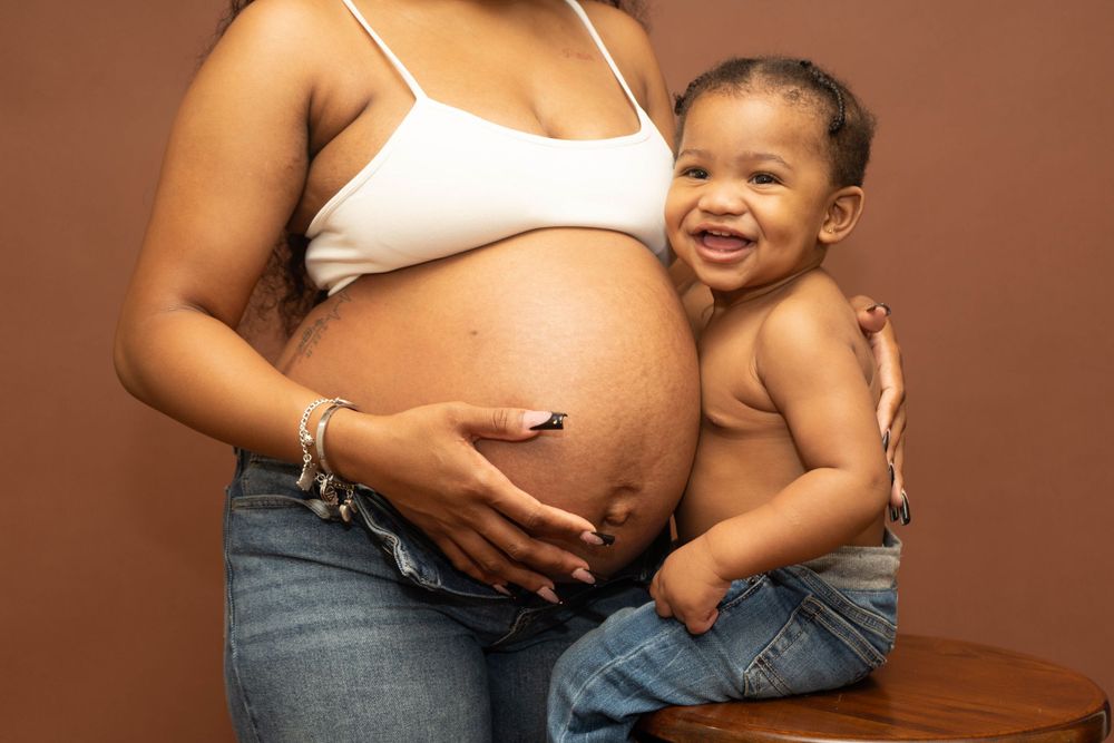 Pregnant woman in white crop top and jeans holds baby boy close to her bare belly during maternity photo session.