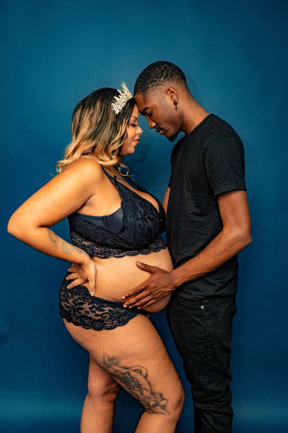Pregnant couple in blue studio, man in black holds baby bump, woman wears navy lace maternity lingerie and crown.