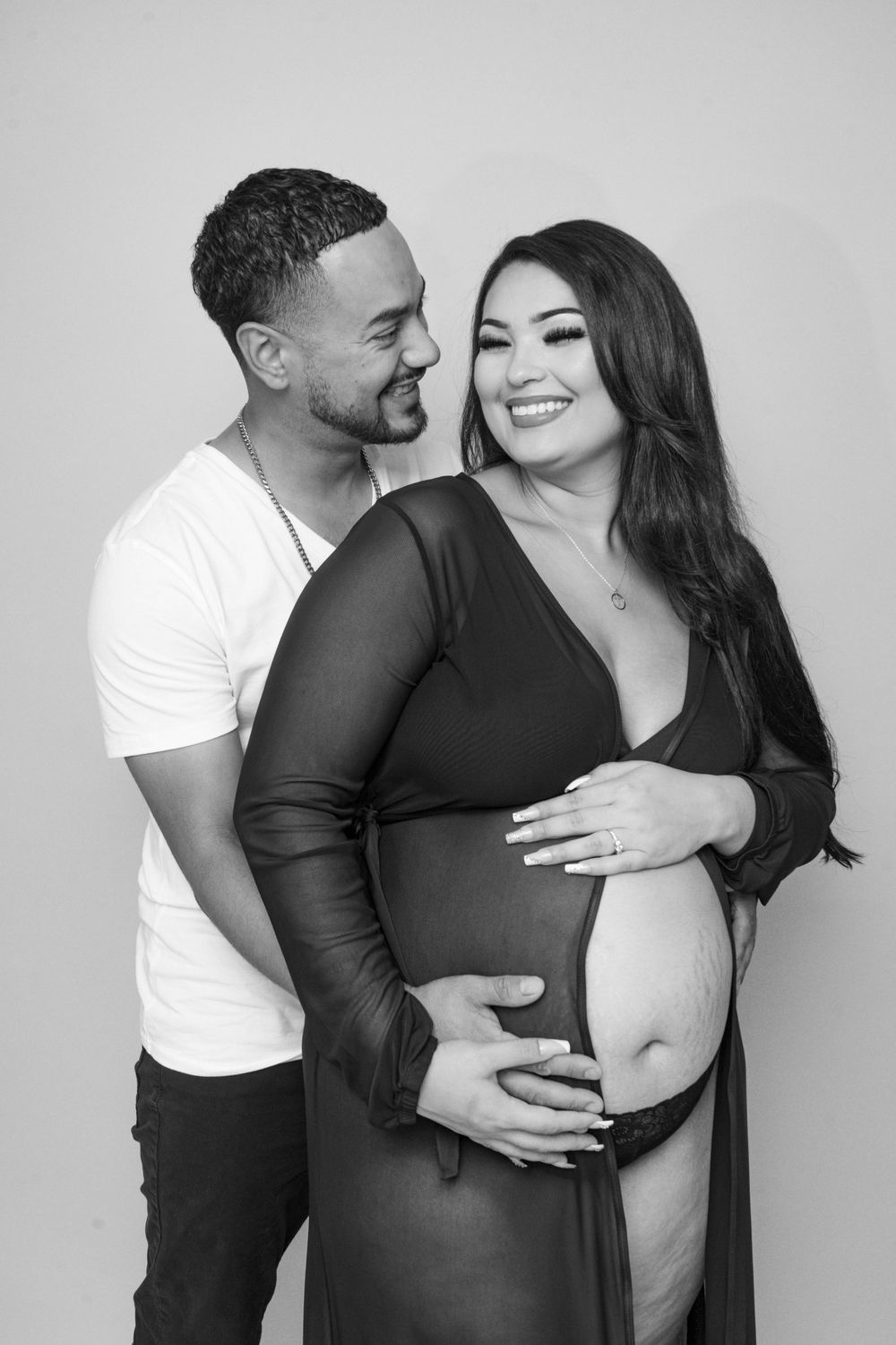 Black and white maternity photo of a smiling couple embracing and cradling a pregnant belly.