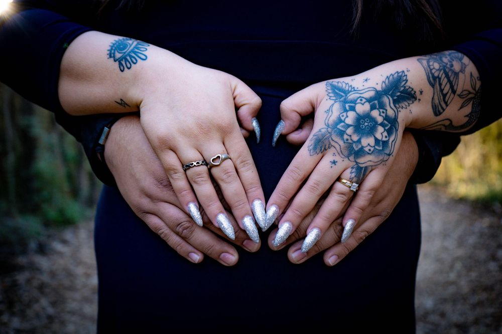 Two pairs of tattooed hands form heart shape on pregnant belly during maternity photo shoot.