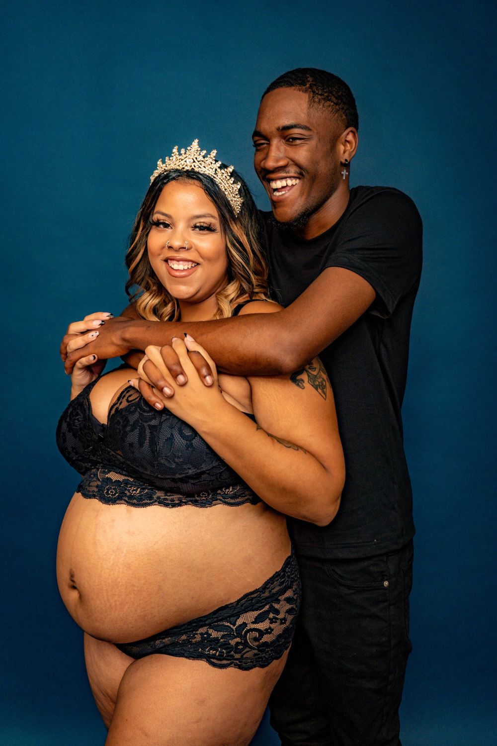 Joyful expecting couple embracing in studio, woman in navy lace lingerie wearing tiara crown.