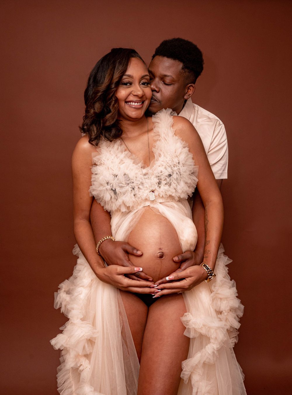 Couple shares tender maternity photo, man kisses woman's cheek as they cradle her baby bump in cream feathered gown.