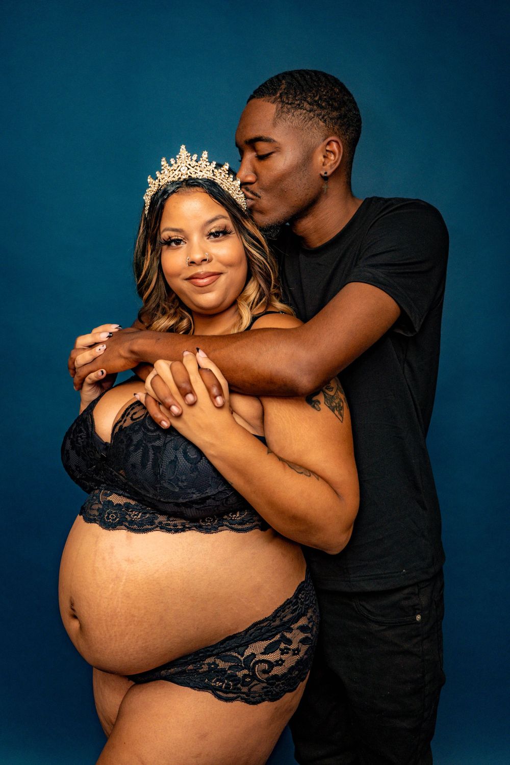 Pregnant woman in navy lace lingerie with crown receives kiss from partner in studio.