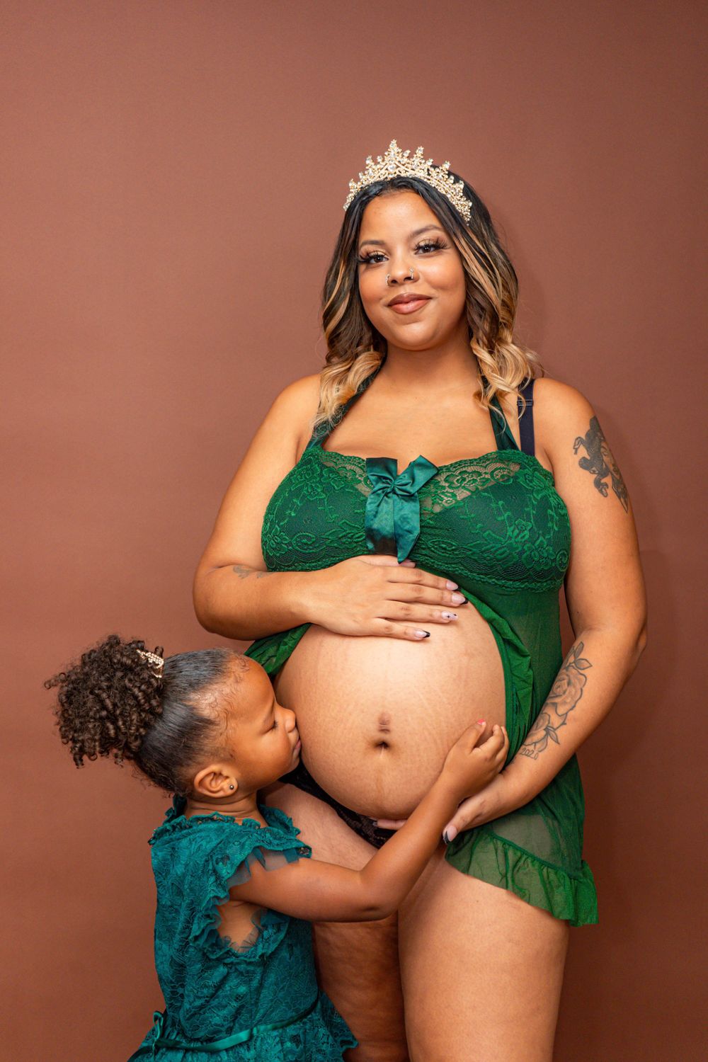 Pregnant woman in green lace lingerie with crown poses with young daughter in studio.