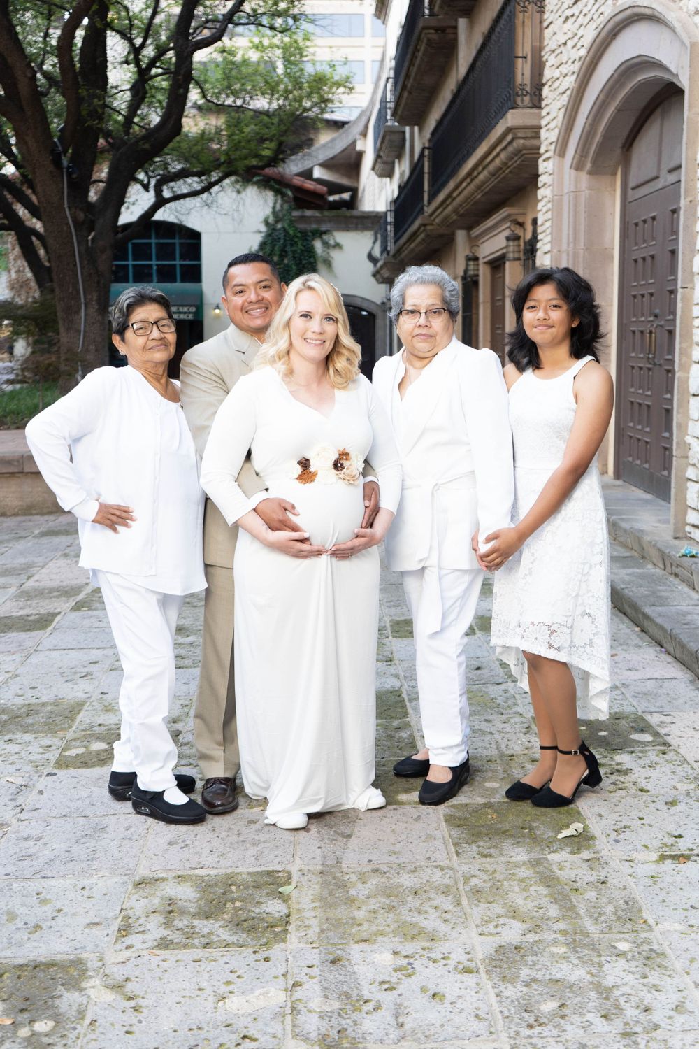 Pregnant blonde woman in white gown poses with family group outdoors for maternity photo session.