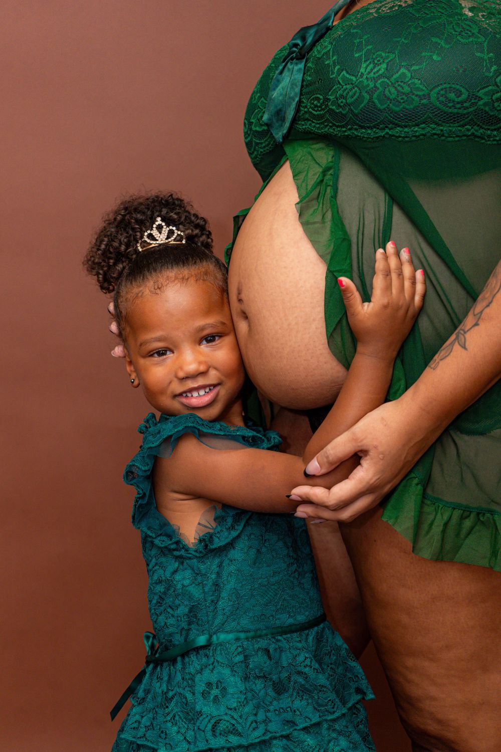 Smiling young girl in teal dress hugging pregnant mothers belly in studio photo.
