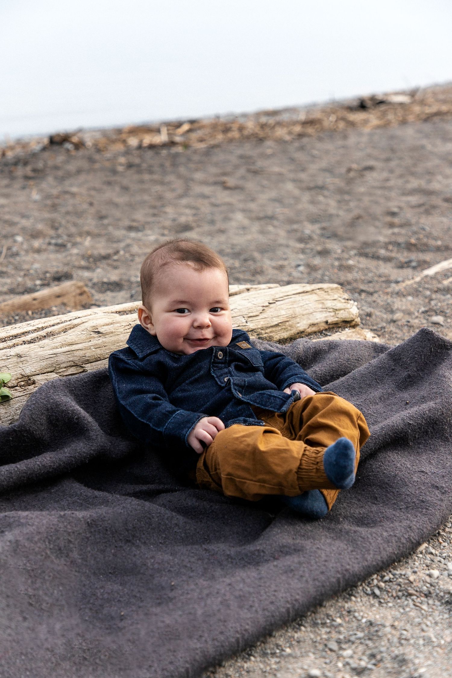 A baby boy sitting on a blanket next to a log at the beach with a cute smirk