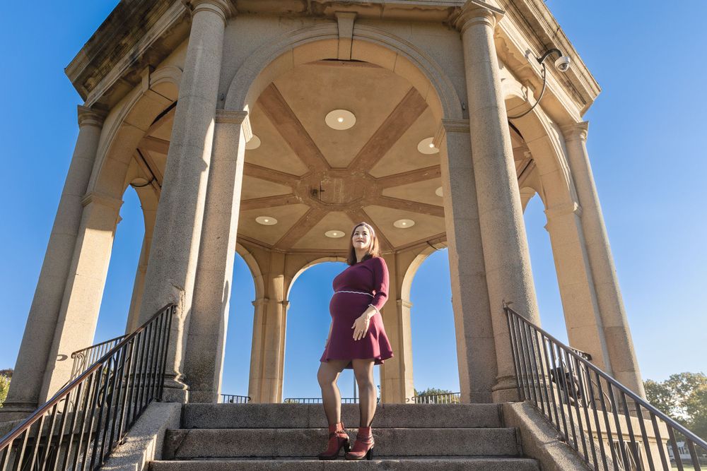 Woman in burgundy dress poses in ornate stone gazebo against blue sky.