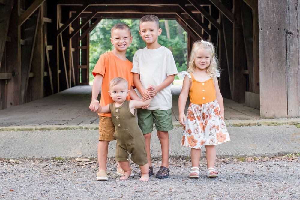 Four young siblings posing together under covered bridge on gravel path.