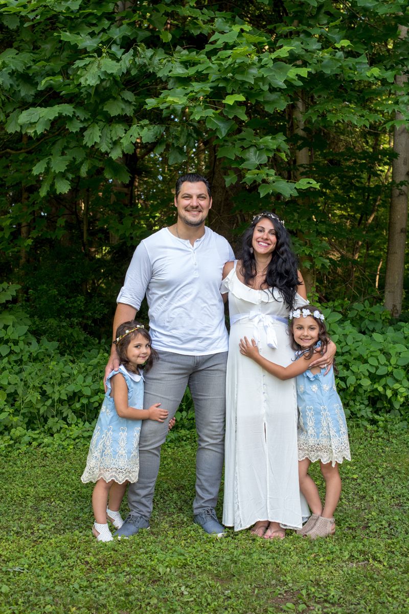 Family of four with pregnant mother and two daughters posing in lush green forest.