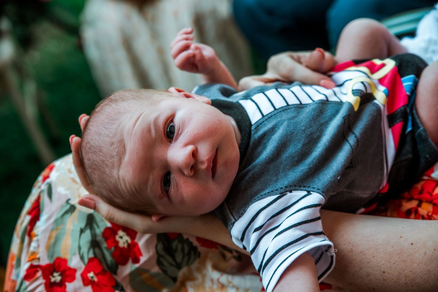 A baby wearing a dark shirt lying on a colorful floral blanket looking at the camera.