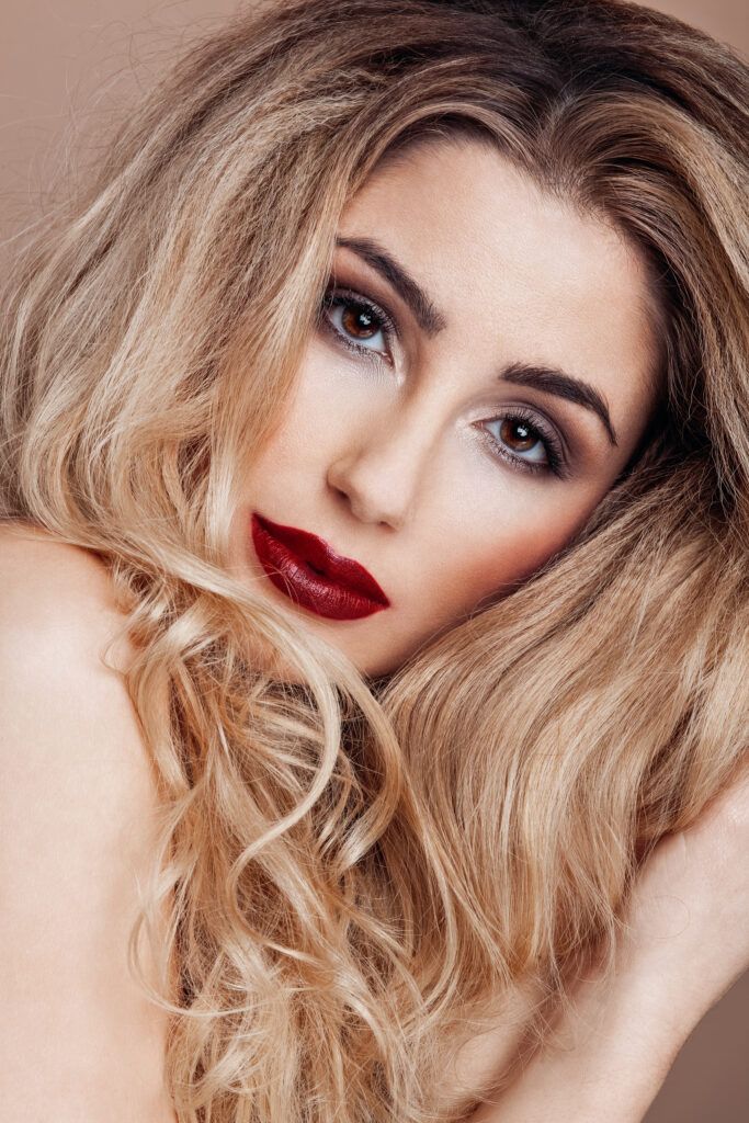 Beauty portrait with wavy blonde hair, dramatic red lips and smokey eye makeup against light background.