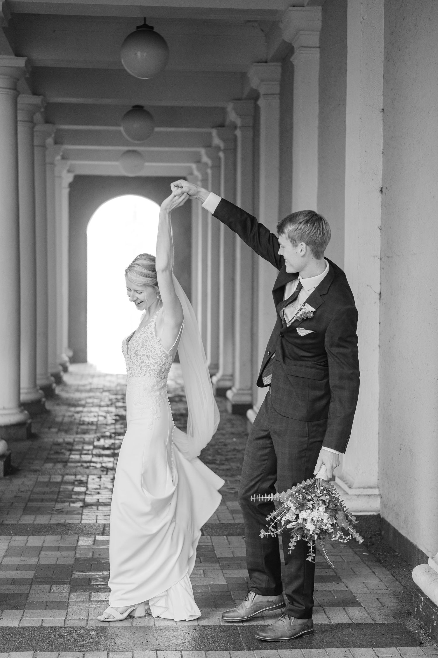Bride and groom pose in elegant black and white photo under arched colonnade.