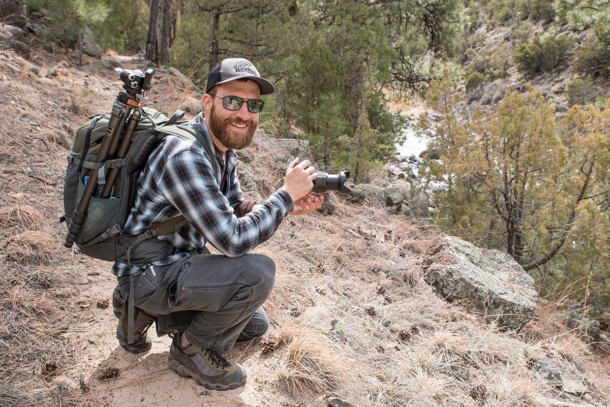 Arkansas commercial and nature photographer, Alex Kent crouches with a camera in hand on a cliff with river below.