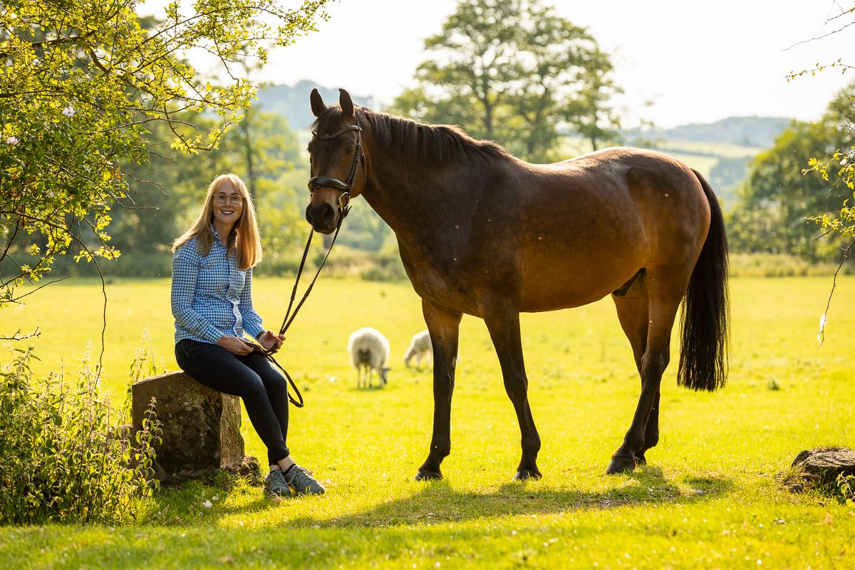 A horse stands beside someone seated on a tree stump in golden sunset light.