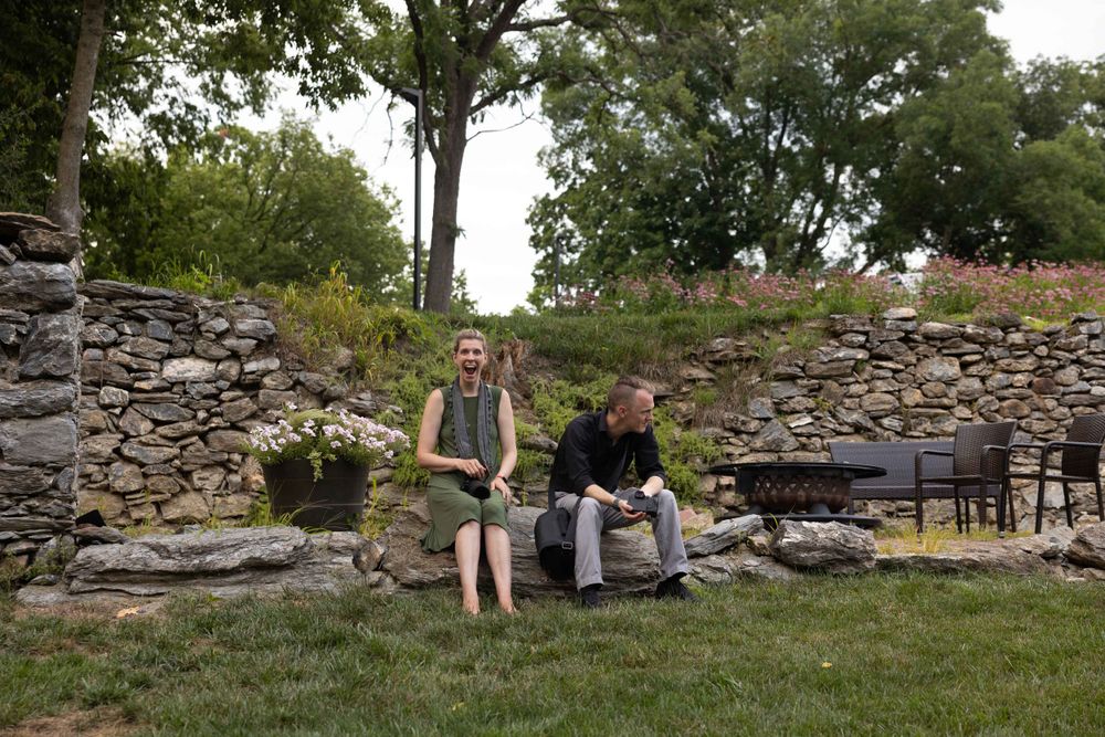 Two people sitting on stone wall with garden flowers and trees in background.