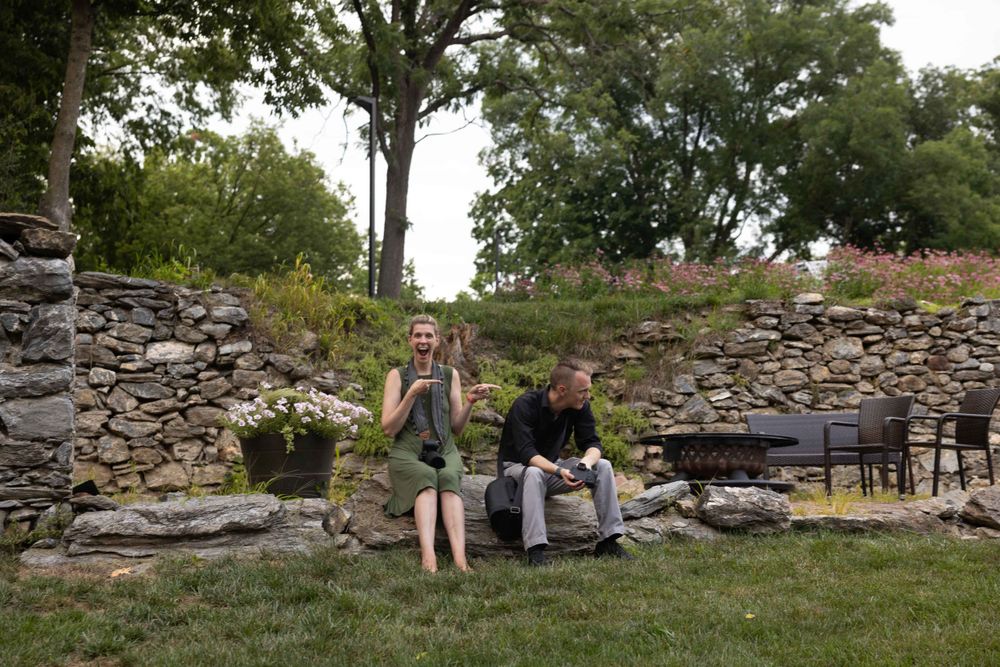 Two people posing casually by stone wall outdoors.