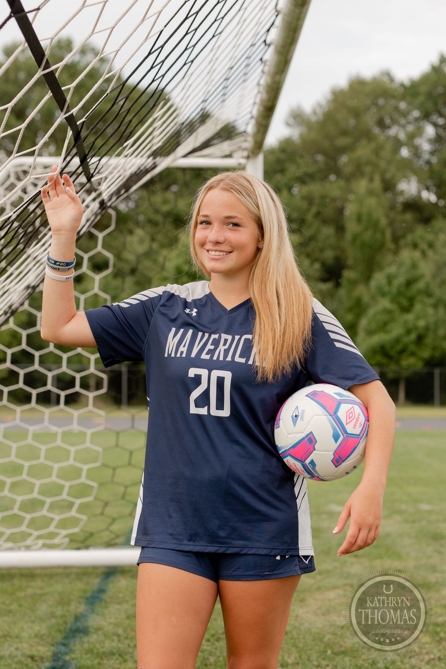Soccer player wearing navy Maverick jersey number 20 holding ball by goal net.