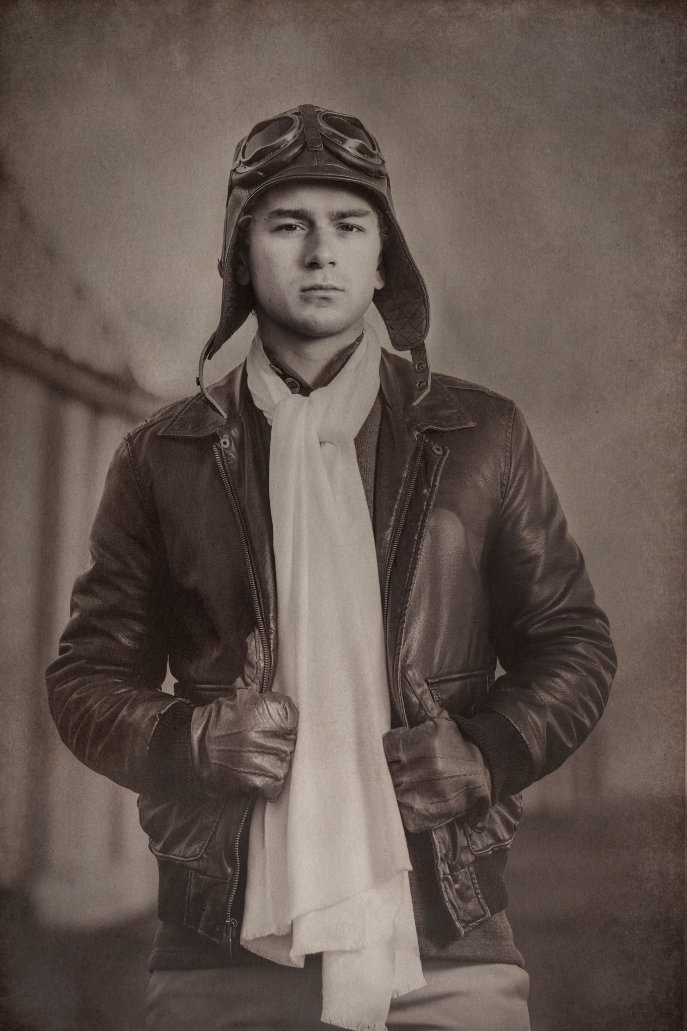 Vintage-style portrait of aviator wearing leather flight jacket and goggles against sepia backdrop.