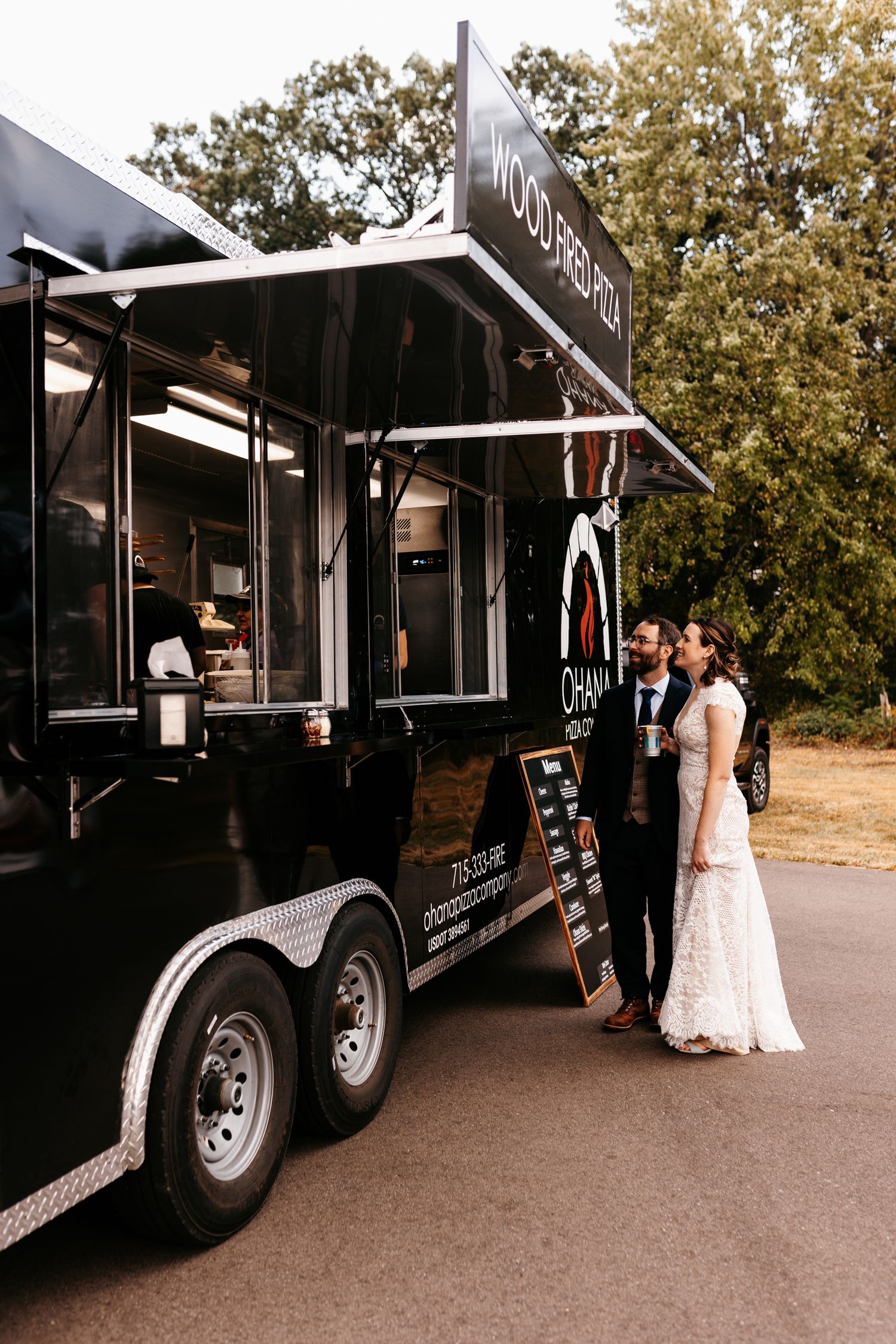 A black food truck serving a couple at a wedding with trees in the background.