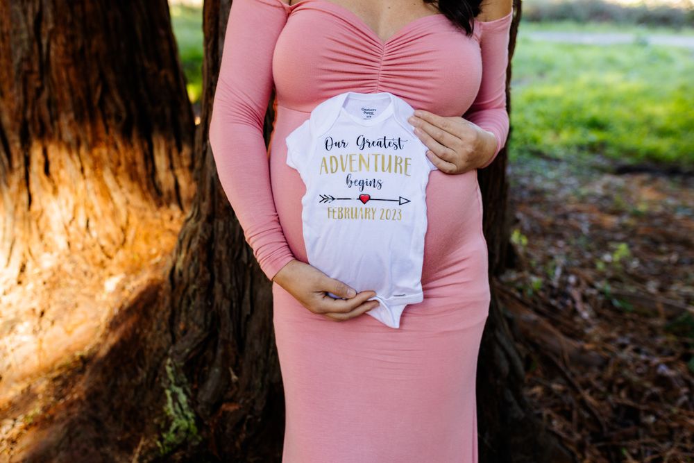 Maternity photo showing a pink dress and baby onesie held against a pregnant belly in an outdoor setting.
