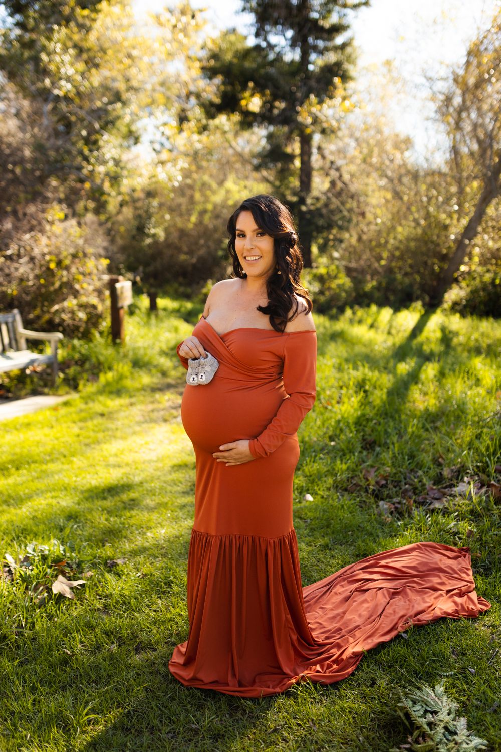 Elegant maternity photo in a rust-colored dress against a sunlit natural backdrop.