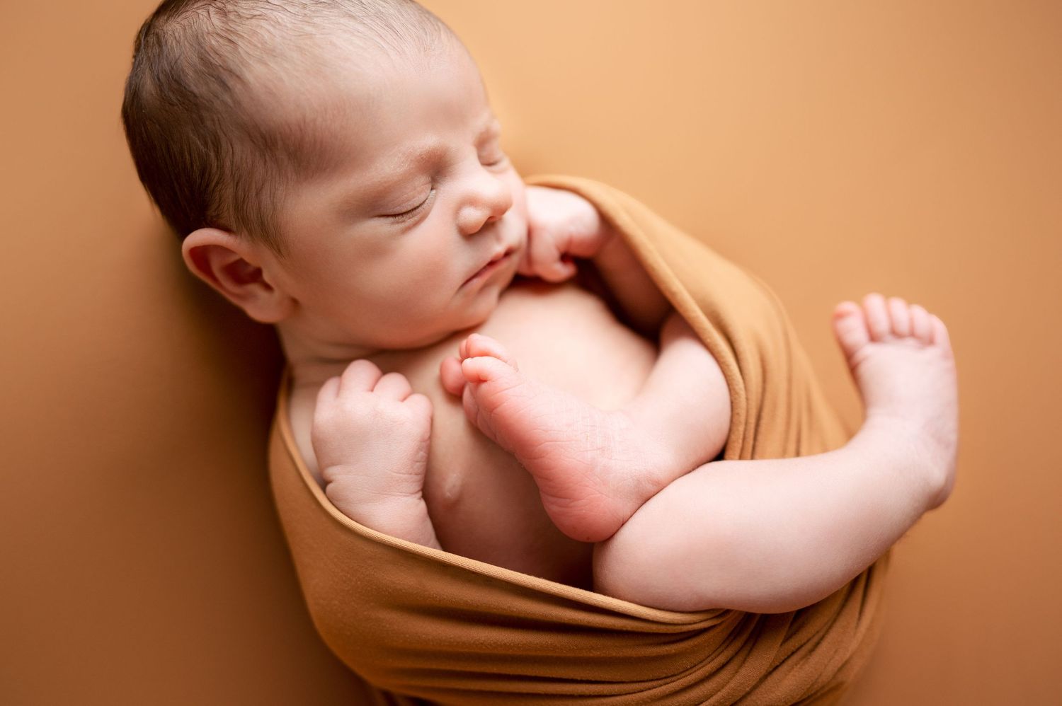 Newborn baby peacefully sleeping in tan colored wrap on brown background.