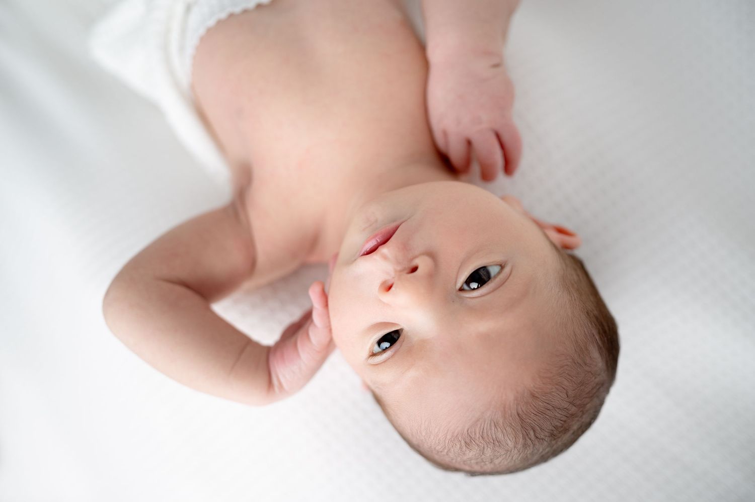 Overhead view of alert newborn baby in white outfit on white background.