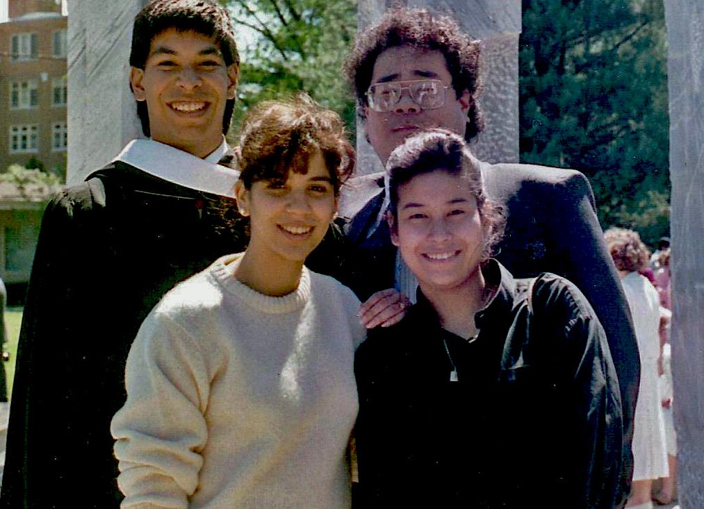 Group of graduates in casual and formal attire smiling together outdoors on campus during graduation day.
