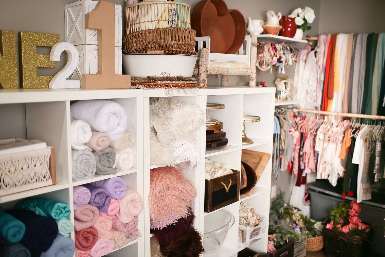 Organized newborn prop storage displays colorful yarn, fabric rolls, and crafting supplies in white cubby shelves.